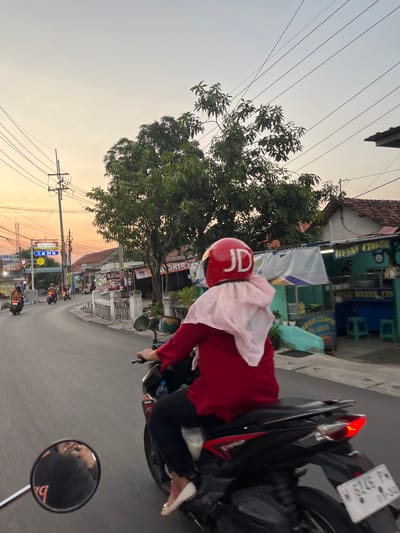 A person wearing a red jacket and a red helmet with "JD" written on it is riding a motorcycle down a street. The rider is also wearing a light pink headscarf. The motorcycle has a license plate that reads "W 6246 PM". A mirror on the motorcycle reflects the rider's face and a pink scarf. In the background, there are buildings with signs in Indonesian, including "Indomaret" and "SATE MAS ROMA". A large tree is also visible, with its leaves casting shadows. The sky is a pale orange, suggesting it is either sunrise or sunset. Overhead power lines run across the scene. The overall impression is of a person commuting or traveling in a tropical or sub-tropical urban environment.