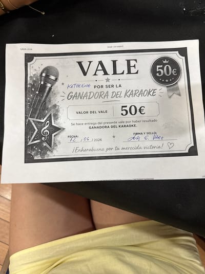 The image shows a "VALE" (voucher) for "GANADORA DEL KARAOKE" (Karaoke Winner). The voucher is for 50€. The date is written as 12/06/2026 and the signature and seal area has handwritten text. The voucher is printed on paper and is the central subject of the image. The voucher is placed on a dark textured surface, possibly a bag or a mat. A small portion of a person's legs wearing yellow clothing is visible at the bottom of the image. The lighting suggests an indoor setting. No specific location in Torrejón de Ardoz, Spain, can be determined from the image.