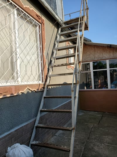 An outdoor image shows a rusty, white metal staircase leading up the side of a building with a gray stucco exterior and a brown textured trim around a window. The window has a white diamond-patterned grate. Adjacent to this is a window with a light brown frame, reflecting trees and a blue sky. A section of the building is covered with light brown plywood. The ground is paved with cracked concrete slabs. A white sack is visible at the base of the wall, near a section of dark brown brickwork. The sky is clear and blue. The scene appears to be in Konstantinovka, Moldova.