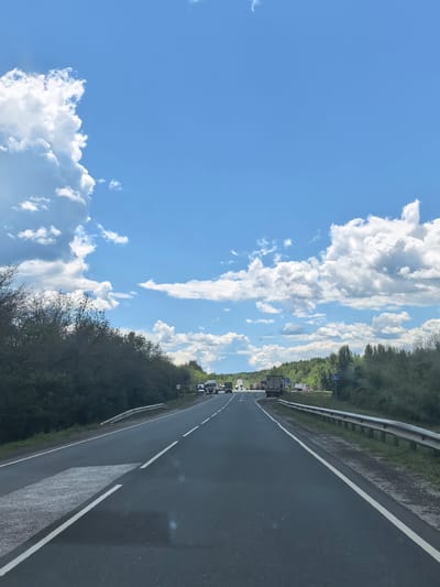 The image captures a daytime view of a highway, likely near Vozrozhdenie, Russia, characterized by a bright blue sky with scattered white clouds. The road is asphalt and appears to be a multi-lane highway, with clear white lane markings. On the left side of the road, there are dense trees and vegetation. On the right side, the landscape transitions to a less dense treeline, with a metal guardrail separating the road from the embankment. Several trucks are visible on the highway, suggesting ongoing traffic. The overall scene depicts a typical highway environment under a sunny sky, with natural elements dominating the periphery.