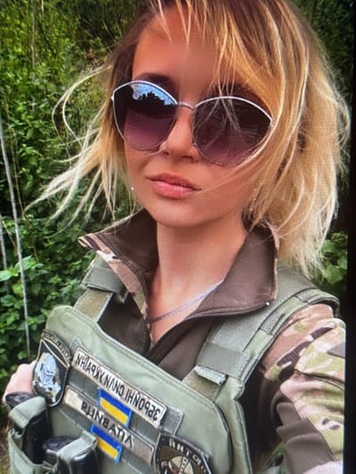 A woman wearing sunglasses and a tactical vest stands in front of a green, leafy background. The vest has several patches, including one with the Ukrainian flag. The woman has blonde hair and is looking towards the camera.