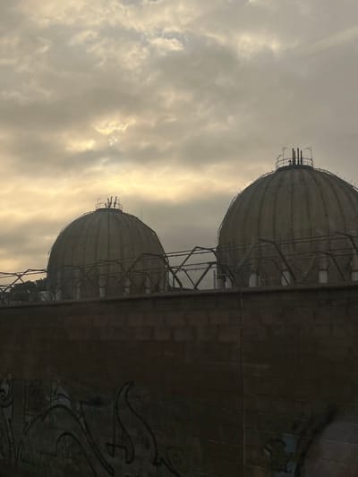 The image displays two large, spherical industrial structures, likely gasometers or storage tanks, under an overcast sky. Both structures are predominantly gray, exhibiting a segmented or ribbed surface texture and intricate metal support frameworks around their upper sections and bases.
The more prominent structure in the foreground is partially framed on its right by a dark, flat industrial wall or building section. On this wall, indistinct, dark graffiti is visible, though specific words or symbols cannot be deciphered.
The sky, which forms the backdrop, is heavily clouded with varying textures. Patches of warm, diffused light break through the clouds, suggesting either the soft illumination of dawn or dusk. The overall lighting is subdued and even. There are no human figures or discernible activity present, emphasizing the monumental scale of the industrial architecture. The scene is situated in Montornès del Vallès, Spain.