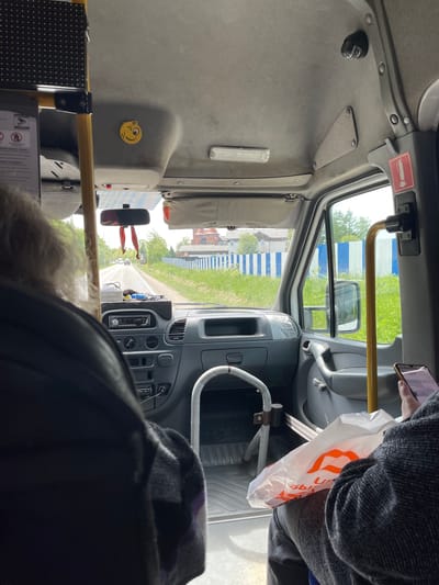 A view from inside a bus on a road in Орловка, Russia. A building with a red roof is visible through the front window. A person is visible on the right side of the image, holding a smartphone and a shopping bag. A steering wheel and dashboard are visible in the center. A handrail is visible. A yellow smiling face sticker is visible on the left.