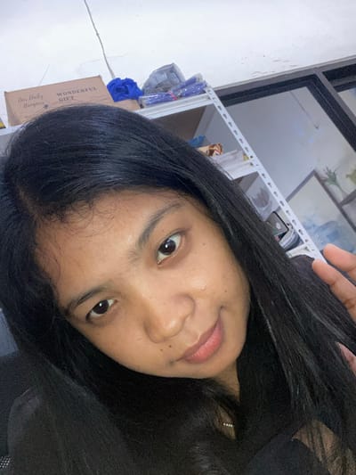 A selfie taken by a woman with long, black hair. The woman's face is the central focus, showing her eyes, nose, and lips. She is wearing an earring. In the background are a window, shelves with various items, a blue fabric, and a cardboard box labeled "Wonderful Gift". The location is Negara, Indonesia.