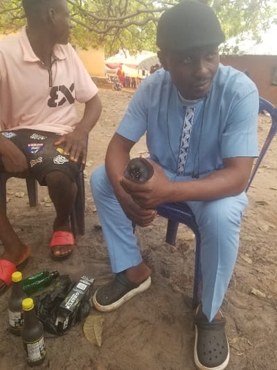 The image depicts an outdoor scene in Ochadam, Nigeria, featuring two male subjects seated on chairs on sandy ground. In the foreground on the right, a dark-skinned male, estimated to be in his 30s-40s, wears a dark flat cap, a light blue short-sleeved shirt with a patterned neckline, light blue trousers, and black clogs with white soles. He is seated on a blue plastic chair, holding a dark, possibly metallic or ceramic, cup or container in both hands, and looking towards the right of the frame. To his left and slightly behind, a second dark-skinned male wears a light pink polo shirt with a black "EX" logo, dark shorts with multiple patches, and red slide sandals. He is seated on a dark chair and faces away from the camera, looking towards the left.
On the sandy ground between the two subjects, two brown plastic bottles with yellow caps are visible, along with a green glass bottle lying on its side. A black plastic bag contains a partially visible white box labeled "GRETUE." Several dried leaves are scattered on the ground. The background reveals an unpaved area with a light-colored building. Further back, under the canopy of a large tree, a red structure resembling a tent or awning is visible, with a red motorcycle partially discernible nearby, suggesting a communal or market area. The scene is illuminated by natural daylight.