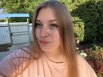 A young woman with blonde hair and blue eyes is taking a selfie outdoors. She is wearing a pink t-shirt and a silver necklace. The background shows a fence, a gate, and lush green trees. The scene appears to be in a residential area or a garden. The image was taken in the city of Sovkhoza "Razdol'e", Russia.