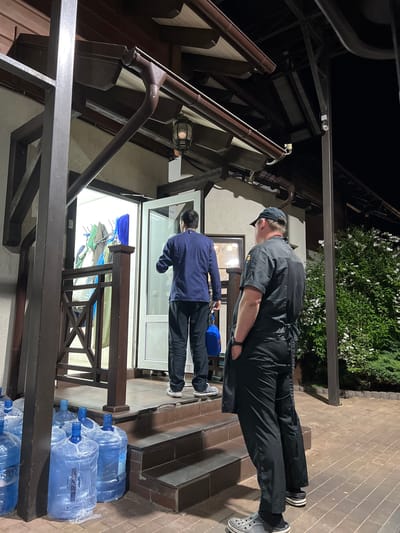 Two individuals are standing outside a building at night. The person on the left is wearing a dark blue jacket and dark pants. This individual is holding a bright blue bag and facing a doorway where several garments are hanging on display. The person on the right is wearing a black chef's uniform and a black baseball cap, with their hands in their pockets, facing away from the camera and towards the right side of the frame. In the foreground, several large blue water jugs are lined up on the ground. The building appears to be a restaurant or similar establishment with an outdoor seating area and a covered entrance. The area is paved with brick. The location is identified as Plastunivska, Russia.