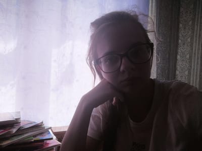The media file is a medium shot, selfie-style photograph taken indoors. The primary subject is a young person, appearing to be female, with light-colored hair pulled up loosely. She is wearing glasses with dark frames and a light-colored t-shirt. Her left arm is propped up, with her hand supporting her chin, and she is looking directly at the camera. Her facial expression appears neutral or slightly pensive.
The background suggests an indoor setting with natural light coming from behind. To the left, a sheer, light-colored curtain with a subtle patterned texture is visible, allowing light to filter through. To the right, a darker curtain with a more ornate, damask-like pattern is present. A stack of colorful books or magazines is piled on a surface in the lower left of the frame. A wooden surface is also visible behind the books.
The lighting creates a silhouette effect on the right side of the subject, while the left side is more illuminated by the light coming through the sheer curtain. This contrast suggests it might be daytime, possibly late afternoon or early morning, depending on the direction of the light. There is no clear indication of the specific location other than the provided context of Dubrovka, Russia. The overall mood is quiet and introspective.
Visible text is present on the subject's t-shirt, appearing as reversed lettering. While partially obscured and in shadow, the letters "DOR" are discernable, suggesting it could be part of a brand name or graphic design.