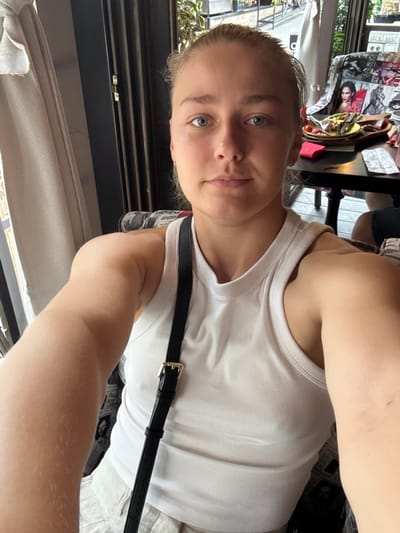 A young woman with blonde hair pulled back and blue eyes is taking a selfie in what appears to be an outdoor cafe. She is wearing a white ribbed tank top and has a black strap from a purse or bag crossing her chest. She is sitting at a table with a plate of food and drinks. In the background, there are chairs covered in magazine pages and some foliage. The scene is well-lit with natural light. There is no information in the image to suggest the location is Predeluț, Romania.
