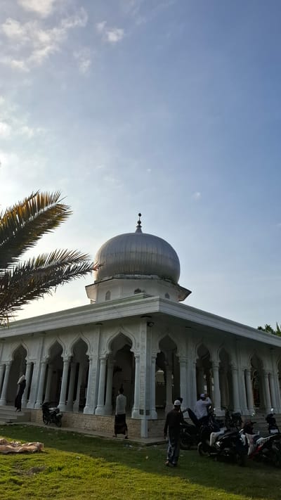An exterior, low-angle, vertical, medium shot captures a mosque in Grong-grong, Indonesia, under a bright, partly cloudy sky. The mosque features a large, silver dome with a finial, perched atop a white, multi-tiered structure. A colonnaded walkway with arched openings surrounds the main building, supported by white pillars.
In the foreground, a well-maintained green lawn covers the ground. Several motorcycles are parked on the grass in front of the colonnade. A person dressed in dark clothing and a white cap stands facing away from the camera near the parked motorcycles. Another person in a light-colored shirt and dark sarong walks to the left, away from the camera. A woman in black is visible walking up steps to the left of the mosque, carrying a white object.
To the left of the mosque, a large palm frond extends into the frame, partially obscuring the sky and the dome. The overall scene is serene, with a sense of community gathering around the religious site. The lighting suggests it is either early morning or late afternoon, with the sun casting a gentle glow.