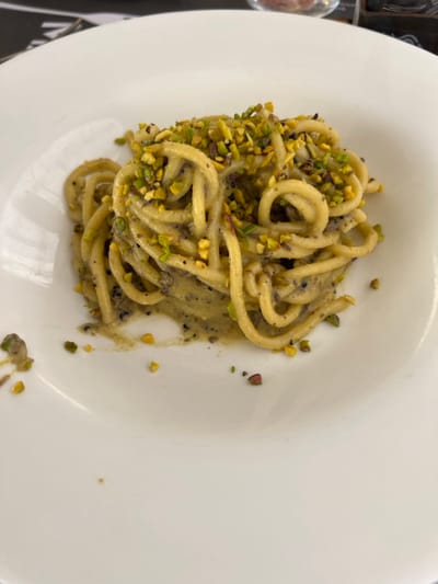 A close-up, top-down view captures a serving of pasta on a white ceramic plate within a dining establishment in Foligno, Italy. The central subject is a mound of thick, light-yellow cylindrical pasta, consistent with bucatini, coated in a light-green, creamy sauce containing dark specks, suggestive of ground truffle or pepper. The pasta is generously topped with roughly chopped green pistachios, some displaying purple-red hues. A few pistachio pieces and traces of sauce are visible on the clean white rim of the plate. The background is blurred, showing a dark surface on the upper left, likely a placemat or table, and a vague, lighter object on the upper right, consistent with another dining item. No individuals are present. The specific address or name of the establishment in Foligno cannot be determined from the visual evidence.