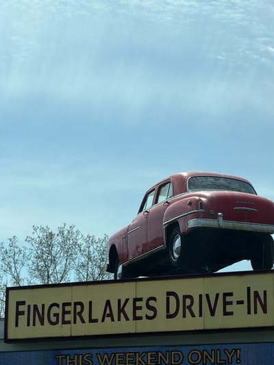 An eye-level shot captures a vintage red car perched atop a sign, set against a bright, partly cloudy blue sky. The car, identified as a Dodge by the emblem on its trunk, appears to be an older model, possibly from the late 1940s or early 1950s, characterized by its rounded design, chrome trim, and hubcaps. Some signs of wear and rust are visible on its body and bumpers.
Below the car, a prominent rectangular sign features the words "FINGERLAKES DRIVE-IN" in dark, bold lettering on a light, aged yellow background. Bare tree branches are visible on the left side of the frame, hinting at the surrounding environment. Below the main sign, a digital or LED display is partially visible, showing the text "THIS WEEKEND ONLY!".
The scene depicts an outdoor setting, likely a commercial establishment given the prominent signage and the displayed car, which serves as an advertising fixture. The bright, clear daylight suggests fair weather. The vintage car and the "DRIVE-IN" text evoke a sense of nostalgia for classic American entertainment venues. The location context of Auburn, United States, aligns with the "Fingerlakes" reference. There are no people visible, and no specific activity other than the static display.