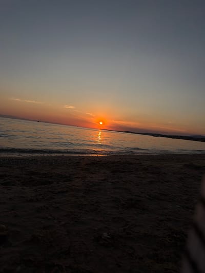 The image depicts a serene sunset over the sea in Πόρτες, Greece. The sky transitions from a soft gray at the top to warm oranges and yellows near the horizon, where the sun is setting. A sliver of the sun is visible, casting a shimmering golden path across the gently rippling water. In the distance, a faint, dark silhouette of landmasses, possibly islands or the mainland coast, is visible along the horizon. The foreground is a sandy beach with scattered pebbles. The overall atmosphere is peaceful and tranquil, characteristic of a sunset by the sea. There are no people or specific objects that could identify the exact location beyond the general description of a beach at sunset.