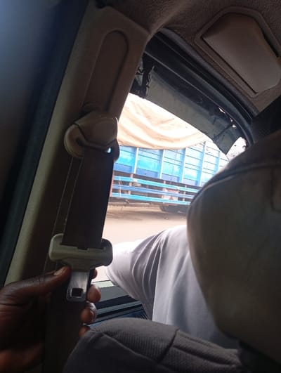 The image captures a scene from inside a vehicle, looking out through the side window. In the immediate foreground, a person's dark-skinned hand is holding the metal buckle of a seatbelt, positioned on the left side of the frame. The seatbelt itself is dark brown or black, running vertically upwards and secured by a light-colored plastic anchor point attached to the car's interior pillar. Part of a vehicle's headrest is visible in the lower right, and the arm and torso of another person, wearing a light-colored t-shirt, are seen seated towards the right, suggesting they are in the front passenger or driver's seat.
Outside the window, under bright daylight conditions, a large truck with a blue frame or body is prominently visible. The truck appears to be carrying cargo covered by a light-colored or beige tarp, possibly indicating goods transportation. The background beyond the truck is somewhat blurry, suggesting a road or ground surface. The car's interior shows a tan or beige ceiling lining and window frame, with some dark material (possibly tape or worn trim) visible around the window edges near the top.
The overall scene suggests a moment during travel, possibly in traffic or stopped by the roadside, as the person holds the seatbelt buckle, perhaps preparing to fasten or unfasten it. The bright lighting indicates it is daytime, likely sunny. There is no visible text. The location context is Tanburawa, Nigeria.