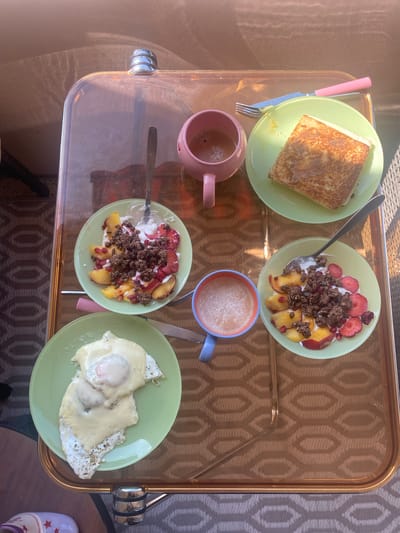 A top-down shot depicts a breakfast spread arranged on a glass-topped tray with a gold-toned patterned design visible beneath. The tray features silver-colored handles. The food items are served on light green plates.

At the top left is a plate of what appears to be toasted bread. Beside it, another plate presents yogurt mixed with fruit (strawberries and peaches) and topped with granola. A mug with a pink exterior contains a beverage.

Directly opposite, at the bottom right, is another plate with two fried eggs, apparently topped with cheese. Next to this plate is another yogurt with fruit and granola. Between the plates is a light blue mug containing a beverage. Cutlery, including forks, knives, and spoons, are placed alongside the plates.

The tray appears to be resting on a gray and white geometric patterned surface.

There is no indication this photo was taken in Monthey, Switzerland. There are no people depicted in the image.
