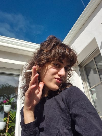 This image depicts a young adult positioned outdoors in Châtelaillon-Plage, France, under a clear blue sky exhibiting a faint contrail. The individual, with brown curly hair and bangs, wears a dark blue long-sleeved garment. They hold a lit white cigarette with an orange filter between the index and middle fingers of their right hand, which is raised with the palm partially visible. The subject is looking directly at the camera. The background features a white building structure, including a visible white gutter system and a multi-paned window on the right side. To the left, a tiered stand supports two white potted plants; one displays pink flowers, and the other contains green foliage. The scene is brightly illuminated by daylight.