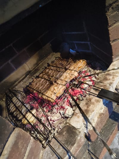 In Востец Снт, Russia, a close-up view shows a grill inside a brick barbecue pit. Two metal grill baskets are positioned over glowing red embers. One basket, on the left, contains what appears to be pieces of white food, possibly chicken or fish. The other basket, larger and positioned in the center and right, holds a substantial piece of raw meat, possibly pork belly or a similar cut, glistening and unseasoned. The meat is laid flat, and the grill bars are visible underneath it. The overall scene captures the initial stages of grilling, with the meat yet to be cooked and the embers providing the heat. The brickwork of the barbecue pit is rustic and appears well-used.