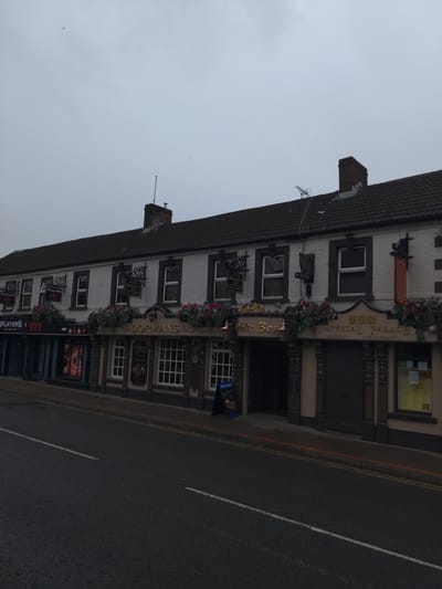 The image shows a row of buildings, including "Dormans" pub and "Imperial Palace Chinese Restaurant" in Magherafelt, United Kingdom, on a cloudy day. The facade of the buildings is white with dark trim and ornate decorations. "Dormans" has a prominent sign with golden lettering and hanging flower baskets. The "Imperial Palace" has a large sign with Chinese characters and "Imperial Palace" in English, also with decorative elements. To the left of "Dormans," a building with a dark facade and signs reading "PLAYERS 777" is visible. The street in front of the buildings is asphalt with white lane markings and a red line along the curb. There are no people visible in the image, and the overall scene is quiet and overcast.