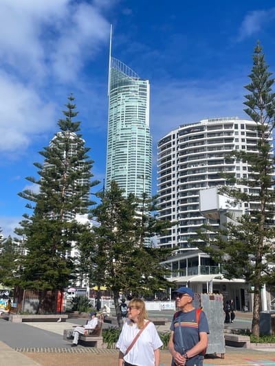 The image captures a daytime scene in Surfers Paradise, Australia, with iconic skyscrapers dominating the background. In the foreground, two people, a woman with blonde hair and glasses wearing a white top and a man wearing a blue cap, a blue t-shirt with stripes, and an orange backpack, are walking. The woman is on the left and the man is on the right. They appear to be walking towards the right of the frame. Behind them, several large Norfolk pine trees stand tall, their distinctive branch structures reaching towards the bright blue sky dotted with white clouds.

Beyond the trees, the distinctive silhouette of the Q1 Tower, a prominent residential skyscraper, is visible on the left. To its right, a curved, white multi-story building, likely a hotel or apartment complex, is also visible. Further to the right, another white residential building with multiple balconies can be seen.

In the mid-ground, there's a paved area with benches, some of which are occupied by other individuals. A small seagull is present on the ground in the mid-left section of the image. The environment suggests a public space, possibly a promenade or park area adjacent to the beach, given the coastal vegetation and the presence of people relaxing outdoors. The overall atmosphere is bright and sunny, indicative of a pleasant day.