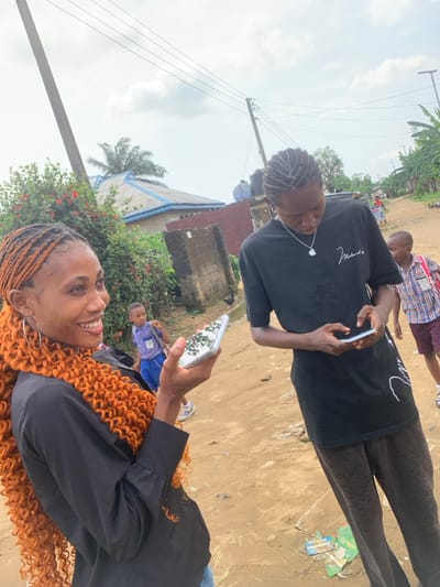 The image depicts an outdoor scene on an unpaved dirt road in the city of Anantigha, Nigeria, under an overcast sky. In the foreground, a young adult female with dark skin, smiling and looking towards the right, is positioned on the left. She wears a black long-sleeved shirt, large silver hoop earrings, and has long, bright orange, tightly curled braided hair extending over her left shoulder. She holds a white smartphone with a reflective, patterned case in her left hand. To her right, a young adult male with dark skin stands, looking down at a white smartphone he holds with both hands. He has braided hair and wears a black short-sleeved t-shirt with a white Apple logo pendant on a silver chain, and dark, patterned pants.
In the background, two young boys are visible. One boy, dressed in a blue and white checkered shirt and blue shorts, carries a blue school bag and is partially obscured behind the female. Another boy, wearing a plaid shirt and maroon shorts, is visible to the right of the male subject. The environment includes a utility pole with overhead power lines on the left, dense green vegetation and bushes with red flowers, and several residential structures, including a building with a light blue corrugated roof and a blue water tank in the distance. The ground shows some scattered debris.