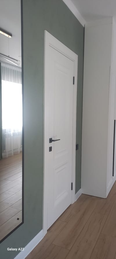 The image depicts an interior scene, likely a hallway or a room with doors. The primary subjects are white doors with dark hardware, including handles and hinges. One door, in the foreground, has two panels and a distinctive L-shaped handle. Another door, further back and higher up, also appears to be white with dark hardware. The walls are painted in two distinct colors: a muted green on the lower section and white on the upper section, which also features part of a white cabinet or built-in furniture. The floor is made of light brown wooden planks laid diagonally. The lighting suggests it is daytime, with natural light illuminating the space from an unseen source, possibly a window or doorway to the right. The overall impression is one of a clean, modern, and well-maintained interior. The text "Galaxy A22" is visible in the lower right corner of the image, indicating the device used for capture. There are no people or animals visible in the scene. The location context is given as Oral, Kazakhstan.