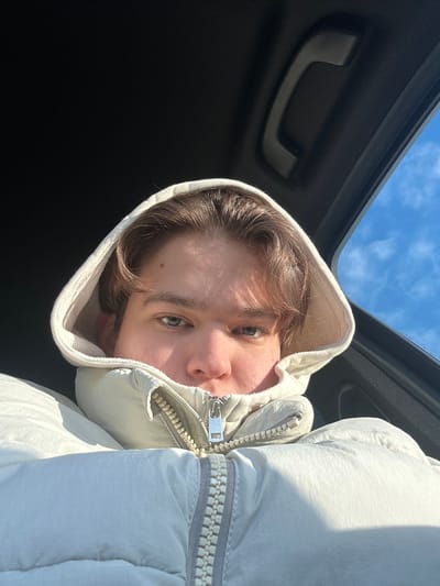 A young person with light brown hair and blue eyes gazes upward from inside a car, wearing a white hooded jacket zipped partially up. The hood frames their face, which is tilted towards the camera. Visible above is a blue sky with light clouds, glimpsed through the car's open sunroof. The interior of the car appears dark. The image is cropped and rotated.