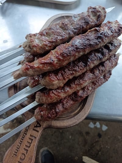 Seven cooked, elongated ground meat kebabs, consistent with lula kebab, are presented on metal skewers, stacked on a dark brown wooden serving board. The kebabs exhibit varying shades of reddish-brown and dark brown, indicative of grilling and charring. Each kebab is threaded onto a flat, metallic skewer, with the exposed skewer ends visible to the left. The wooden serving board features a raised lip and a paddle-shaped handle in the lower-left, engraved with the text "Al Imrol restaurant." The board rests on a reflective, light-grey metallic surface, likely a stainless steel counter or table. Below the serving board, a dark, textured surface is visible, partially obscured by the toe of a dark-colored shoe in the lower-left corner. A blurred, light-colored rectangular object is faintly visible in the upper background. This scene is situated in Kasri Arifan, Uzbekistan.