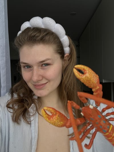 A fair-skinned woman with light brown, wavy hair smiles at the camera. She wears a fluffy, white headband. Positioned near her head is an orange toy lobster. She is wearing a striped shirt. The background consists of a wall on the left and a partially visible window or curtain below.
