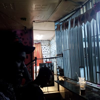 The image shows the interior of a bar in Kinoo ward, Kenya. Two people are visible in silhouette. A partially visible man is in the foreground wearing a hat. A second person is behind him, partially visible, and wearing headphones. Behind them is a bar with metal bars, and a bar top with at least two empty plastic cups. Hanging above the bars is a garland made from playing card suits.
Through a doorway is another room with a red curtain and spotted wallpaper. The ceiling is decorated with ceiling tiles and a hanging light fixture.