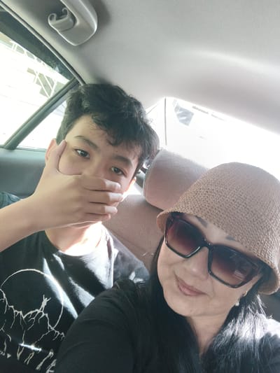 The image shows two people in the back seat of a car. On the left, a young male with dark hair is covering his mouth with his hand. He is wearing a black t-shirt with a white graphic print. On the right, a woman wearing a wide-brimmed hat and large sunglasses is smiling. She has dark hair and is wearing a pearl earring. The interior of the car is visible, including the car's ceiling and headrests. The lighting suggests it is daytime. The image does not contain any specific landmarks that would allow for identification of its location within Yahşihan, Turkey.