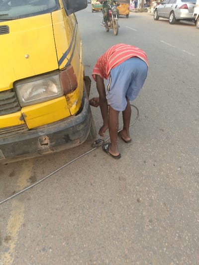 The image captures a daytime street scene in Ijaiye, Nigeria, showing a person attending to a vehicle by the roadside. In the foreground, a person, likely male, is bent over at the front left side of a yellow van, appearing to inflate its tire. He wears a red and white striped short-sleeved shirt, blue shorts, and black flip-flops. A metallic hose or cable extends from a tire pump-like device on the ground, connecting to the van's front tire valve.
The yellow van or minibus, possibly a public transport vehicle, shows signs of use, including a scuffed black front bumper with a visible hole and some damage around the headlight assembly.
In the background, a street with light traffic is visible. A person on a motorcycle is riding away from the viewer, and a yellow and green tricycle (locally known as a Keke Napep) can be seen further down the road. A silver car is parked on the right side of the street. The lighting indicates clear, sunny weather. No visible text is discernible on any prominent objects in the image.