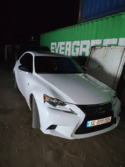A white Lexus sedan is parked on a dark, dirt ground. The car is facing forward and slightly to the right. The front license plate reads "GE SE-099-MO". Behind the car, a bright green shipping container with "EVERGREEN" written on it in white letters is visible. To the right of the car and behind it, a large, light-colored metal door or panel is propped open. The scene appears to be in an industrial or storage area, possibly at night given the lighting. The location is Krtsanisi, Georgia. There are no people clearly visible in the image.