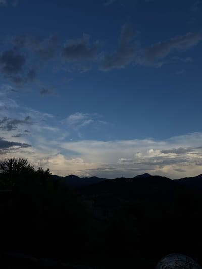 The image captures a landscape under a cloudy sky, transitioning from a clear blue to an overcast horizon. Silhouetted mountains are visible on the right, with dark trees and indistinct structures in the foreground. A pale, round object, possibly the moon, is present in the sky. The location and the city of Zhaltusha, Bulgaria, is implied.