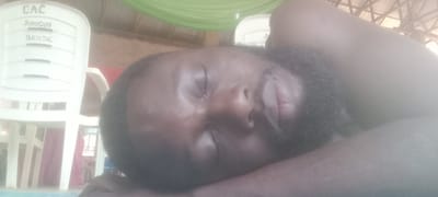 A close-up image shows a Black man with a beard lying down with his eyes closed, possibly sleeping. A white plastic chair with the inscription "CAC AMAOGWU BENDE" is visible in the background. The location is likely Amaogwu, Bende, Nigeria, based on the text on the chair. The surrounding environment appears to be indoors, with green and white fabric or decorations hanging overhead. A metal roof structure is also visible.