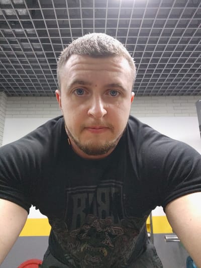 A male individual is depicted in a frontal, chest-up portrait within an indoor gym or fitness facility in Pyt'-Yakh, Russia. The subject has short, light brown hair, blue eyes, and a light brown goatee. He is wearing a dark, short-sleeved t-shirt with a partially visible graphic design on the front, including dark text and an intricate illustration. A thin, light-colored cord or string is visible around his neck. The ceiling features a dark grid structure with integrated light sources. The background includes a white brick wall on the left, and metallic gym equipment along with a grey wall on the right. A red weight plate is partially visible on the dark floor in the lower left foreground. A vertical yellow stripe is visible on a surface in the mid-ground. The subject maintains a neutral facial expression, looking directly at the camera.