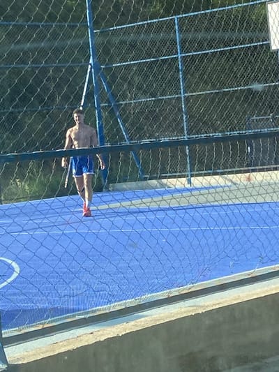 A shirtless man wearing blue and white athletic shorts and red sneakers is walking on a blue sports court. He is holding a white object, possibly a paddle or racket, in his right hand. The court is enclosed by a chain-link fence, and behind the fence, trees and foliage are visible. The overall scene appears to be outdoors on a sunny day. The location is Seoce, Montenegro.