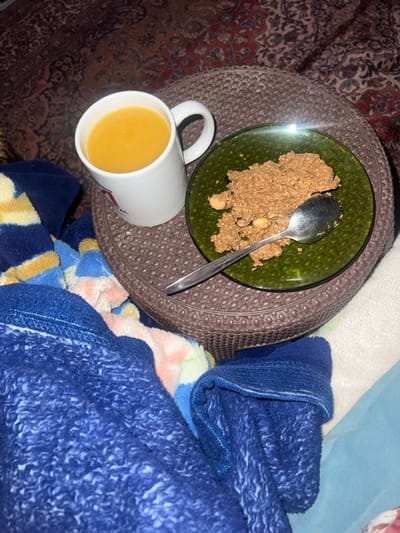 The image presents an indoor scene located in Ksar-el-Kebir, Morocco. A white ceramic mug, filled with a bright orange-yellow liquid, rests on the left side of a round, dark brown, textured side table or tray. To the right of the mug, a dark green glass plate contains a spread of granular, brown food, possibly a type of cereal mixture or dessert, with several light-colored, oval-shaped inclusions resembling nuts. A silver-colored spoon lies diagonally across the food on the plate. The lower portion of the frame is occupied by draped textiles: a dark blue, textured fleece blanket or garment covers the majority of the bottom left, partially obscuring a multi-colored blanket with blue, yellow, and light-colored stripes. A light-colored textile, possibly another blanket, is visible on the far right. The background behind the table shows a patterned rug in reddish-brown and darker tones. No individuals are visible, and the objects are stationary.