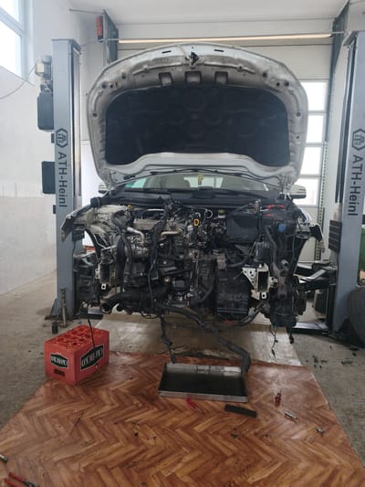 The image displays an automotive repair bay within a garage located in Mrzenci, North Macedonia. A light-colored passenger vehicle is centrally positioned, with its hood fully open and the entire front section, including the bumper and fenders, removed. The vehicle's engine and transmission assembly are exposed and appear partially disconnected or lowered within the engine bay. The car is situated between two grey two-post vehicle lifts, both branded "ATH-Heinl."

Beneath the vehicle, a rectangular sheet of wood-patterned material, resembling herringbone parquet, covers a section of the concrete floor. On this patterned surface, a red plastic crate, bearing the "СКОПСКО" (Skopsko) logo, rests to the left of the vehicle's centerline. A rectangular metal tray, containing a dark liquid consistent with engine oil or coolant, is centrally placed on the patterned sheet directly beneath the engine area. Several hand tools, including red-handled screwdrivers, socket wrench components, and pliers, are scattered on the patterned sheet and the adjacent concrete floor. The garage walls feature white tiling in the lower section and plain white paint above. Natural light enters through large windows or translucent panels visible in the background, supplemented by overhead fluorescent lighting. A single tire is visible on the concrete floor near the base of the right vehicle lift. No individuals are present in the scene.