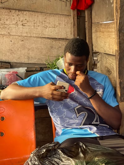 The image displays a young man in what appears to be a rustic or makeshift indoor setting, captured in a moment of quiet contemplation or focus. The original image is oriented sideways (rotated 90 degrees counter-clockwise), but the description will assume an upright perspective.
The central figure is a young man with dark skin and short, dark, curly hair. He is wearing a light blue short-sleeved t-shirt or sports jersey, which has a darker blue pattern (possibly stylized shapes or leaves) across the chest and shoulders, and a large white number "12" prominently displayed on the front. The collar is dark blue with a thin yellow stripe. On his left wrist, he wears a black beaded bracelet.
He is holding a light-colored smartphone in his right hand. The phone is encased in a distinctive white cover featuring a red ice cream cone design on its upper part and a simple, drawn-on cartoon face (two dots for eyes, a curved line for a mouth) on its body. His gaze is directed downwards and slightly to his right, suggesting he is looking at something off-frame or intently at his phone. His left hand is raised, with fingers slightly curled, resting near his chin or mouth, conveying a pensive or relaxed posture.
The background consists primarily of rough, untreated wooden planks, forming what appears to be a wall or part of a simple structure. The wood is light-colored and shows visible grain and texture, with some gaps between the planks.
Several other objects are visible in the scene:
* An orange plastic stool or chair occupies the lower right foreground, with the man's right forearm resting on it.
* To the right of the man, a crumpled black plastic bag is visible.
* Behind him, to his left, is a translucent white plastic container holding some green leafy vegetation, possibly herbs or vegetables.
* Further to the left, a red fabric item, possibly a piece of clothing, is partially visible against the wooden wall.
* Another clear plastic container is visible on the ground or a lower surface beneath the green vegetation.
The lighting suggests it is daytime, with natural light entering from the right side of the frame (when upright), casting subtle shadows and highlighting parts of the man's face and arms. The overall atmosphere is unpretentious and casual, focusing on the man's absorbed state. No specific text is clearly legible other than the number "12" on his jersey and the simple face on the phone case.