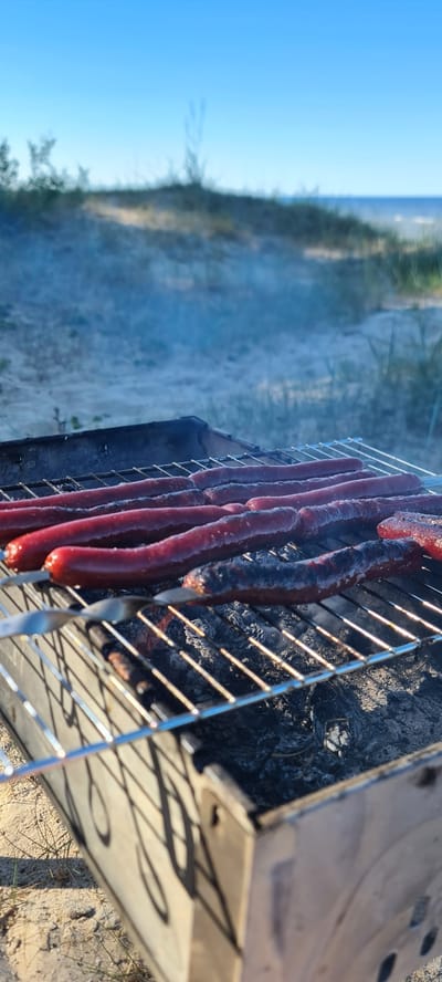 The image captures a portable metal grill in operation, actively cooking multiple long, reddish sausages. The sausages are arranged on a metal grate, with several impaled on skewers. One sausage in the foreground displays significant charring on its surface. Smoke rises from the burning coals or charcoal contained within the grill. The grill itself is a utilitarian, rectangular metal structure. It rests on sandy, light-colored ground with scattered sparse vegetation, primarily dry grasses. In the midground, a low sandy dune or rise is visible, also covered with sparse vegetation. The background features a clear, bright blue sky extending above a calm body of water, likely the sea or a large lake, with a distinct horizon line. The scene is illuminated by natural sunlight, casting long shadows from the grill grate onto the grill's side and the ground. No individuals are present in the frame. The location is Zīvartiņš, Latvia.