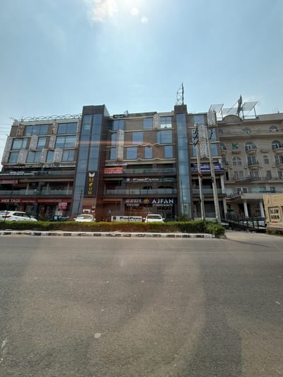 This is a daytime, eye-level, outdoor shot of a modern commercial building in Zirakpur, India. The building features a facade of glass windows and concrete with decorative metal elements. Various businesses are advertised on the building's exterior with visible signage, including "AJFAN DATES & NUTS," "FINE FERN STUDIO," "HERITAGE ETHNIC WEAR," and "Regus." There are also signs for "HAND FOR HUMANITY" and "AKHIL GUPTA ARCHITECT & INTERIOR DESIGNER." A street runs alongside the building, with a white car parked on the side. The sky is clear and bright, suggesting sunny weather. The overall scene depicts a bustling commercial area.