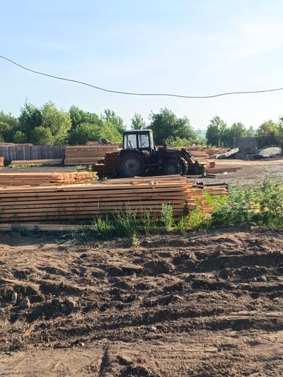 A daytime, eye-level, full outdoor shot captures a lumber yard with stacks of lumber and a tractor. The scene is set against a clear, light blue sky with some wispy clouds, and a line of trees forms the background. In the foreground, dirt with tire tracks is visible, suggesting recent vehicle activity.
Dominating the mid-ground is a large pile of light-colored wooden planks, neatly stacked horizontally. To the right of these planks, a dark-colored tractor with a front loader is parked. The tractor's front tires are large and have visible tread. Additional stacks of lumber are present behind the tractor, some appearing more weathered than the planks in the foreground. A wooden fence runs horizontally across the middle ground, separating the lumber yard from the treeline. The trees are lush and green, indicating a summer or late spring setting. The overall impression is of an active lumber processing or storage facility. The location is identified as Strelka, Russia.