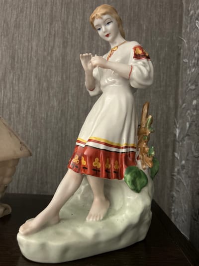 A detailed, forensic, matter-of-fact description of the image:
The image displays a porcelain figurine of a young woman in traditional folk attire. The figurine is positioned on a light green, textured base, suggesting a naturalistic setting like a rocky outcrop or earth. The woman has fair skin, blonde hair styled with a slight wave, and striking blue eyes with dramatic black eyeliner. Her lips are painted a bold red.
She is depicted wearing a white, long-sleeved dress with intricate red and yellow floral patterns around the hem and on the cuffs. The neckline features a yellow trim and a decorative pendant. Her left leg is extended forward, with her left foot bare and resting on the base, while her right leg is bent, and her right foot is also bare and visible. Her hands are held up near her chest, with fingers slightly curled, possibly in a gesture of playing an instrument or performing a dance.
Behind the figurine, a small, brown, twig-like structure with green leaf elements rises from the base. The background consists of a textured gray wall with vertical lines, likely wallpaper, and a hint of a lighter, patterned fabric on the right side, possibly curtains. To the left of the figurine, a portion of another object is visible, appearing to be a light-colored, possibly marble, decorative element with a conical shape.
The overall scene suggests an indoor display setting, with the figurine positioned on a dark, flat surface, likely a shelf or table. There is no discernible connection to the city of Лосева, Belarus.