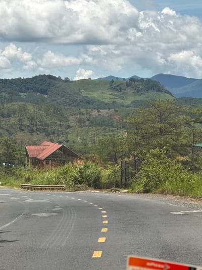 The image shows a paved road curving to the right, with yellow dashed lines marking the lane. On the left side of the road, there is a weathered wooden guardrail. Beyond the road, a dense landscape unfolds with lush green vegetation, including tall grasses and trees. In the middle ground, a partially constructed building with a red tiled roof stands amidst the greenery. The background features rolling hills covered in trees and distant mountains under a sky filled with scattered clouds. The scene is likely located in a rural area of Tu Póh, Vietnam, characterized by its natural beauty and mountainous terrain.
