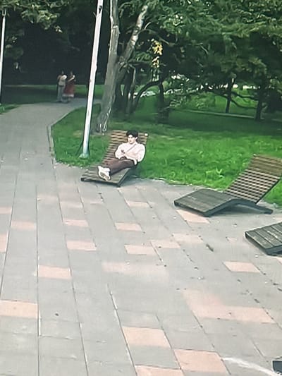 The image depicts an outdoor scene in a park-like setting. In the foreground, a paved walkway with rectangular, light-colored tiles interspersed with darker ones leads into the midground. To the right of the walkway, several dark, slatted chaise lounges are visible on the pavement. Further back, a young person with dark hair is seated on one of these chaise lounges, wearing a light-colored shirt and dark pants. Their arms are resting on their lap, and they appear to be looking downwards. Behind the chaise lounge and the young person, a grassy area is present, bordered by trees with dense green foliage. In the far background, on a path winding through the greenery, two figures, one in a red dress and another in lighter clothing, are walking away from the camera. The overall lighting suggests it is daytime. The scene is likely a public park or recreational area.