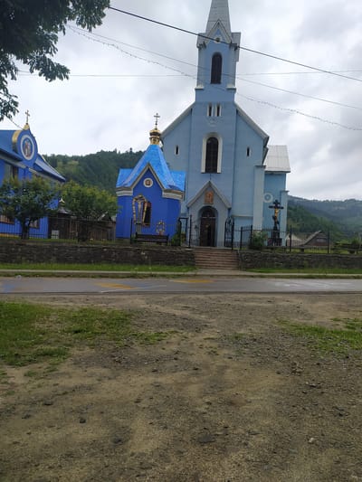 The image shows a bright blue church with a tall steeple and a smaller, similarly colored chapel. The church has a stone foundation and a fenced-in area with a cross on a pedestal in front of it. The chapel has a pointed roof and a small entrance. In the background, there are green hills and a cloudy sky. To the left of the church is another blue building with a golden dome, possibly part of the same complex. The scene appears to be in a rural area, with a road in the foreground and a patch of grass and dirt. The overall impression is of a serene and picturesque religious site.
