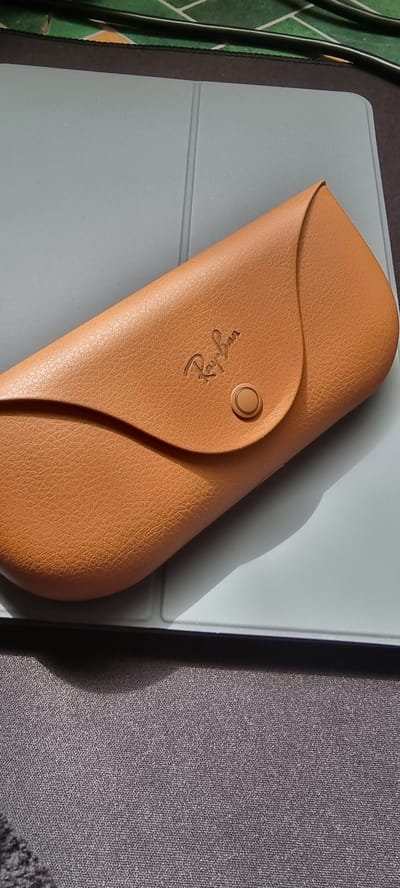 A tan faux leather glasses case with a gold-colored snap closure and the embossed logo "Ray-Ban" is placed diagonally on a light gray surface, possibly a tablet or laptop cover. The case is positioned such that the snap closure is visible on the right side. The background consists of a dark gray textured surface. The scene is brightly lit, casting a shadow from the glasses case onto the gray surface. The image does not provide any information related to the city of Naaldwijk, Netherlands.