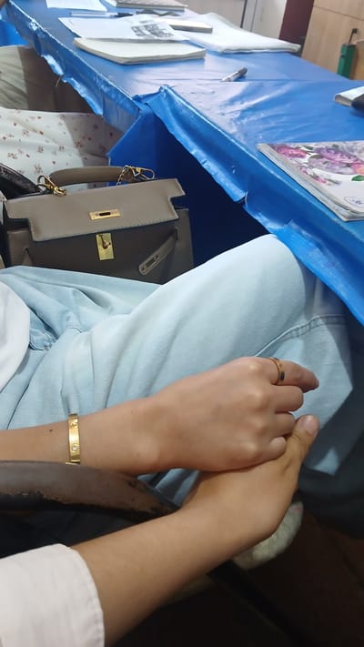 The image depicts a close-up of a person's lap and arms, suggesting they are seated. The individual is wearing light blue denim jeans and a white shirt. On their left wrist, there is a gold bracelet adorned with what appear to be diamonds. A gold ring is visible on the right ring finger.
In the background, a light brown handbag with gold hardware rests on a surface covered by a blue tarp. The blue tarp is draped over a table, on which several items are placed, including papers and what looks like a magazine with floral imagery. The environment appears to be indoors, potentially a room or a space set up for an event or meeting, given the presence of the table and tarp. The overall lighting suggests it is daytime. No specific landmarks or architectural features are visible that would indicate a precise location within Kattakurgan City, Uzbekistan. The image focuses primarily on the personal accessories and clothing of the individual.
