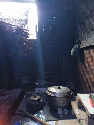 This image captures an interior scene in Na Sangkham, Laos, characterized by low light with a prominent external light source. A strong, diagonal beam of light enters from an opening in the upper left, illuminating airborne particles. To the upper left, a stack of cut wood or timber is piled. The right wall is constructed from woven material, likely bamboo, with a horizontal wooden beam from which a dark green bag hangs. On the earthen floor in the center, a dark, lidded pot sits to the left of a larger, metallic two-tiered steamer pot, which rests on several stones. The lower left foreground contains a wooden stick, clear plastic bags, and a faint orange glow from an unidentifiable source. No individuals are present within the frame.