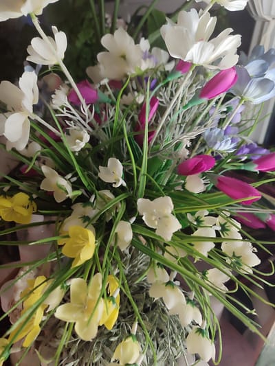 The image shows a close-up view of an artificial flower arrangement. Dominant colors include white, yellow, pink, and green. There are various types of artificial flowers, with small white ones and larger, brighter yellow flowers clustered towards the bottom. Sprigs of thin, green grass-like material protrude upwards between the flowers. Other flowers are of varying colors including white, pink, blue, and purple. The flowers are arranged in a container, which is visible only at the bottom of the image. The location is unknown. There is no indication that this image was taken in Kotlas, Russia.